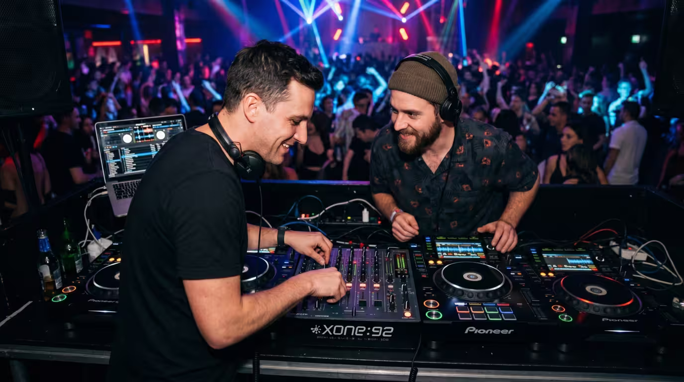 Two DJs sharing a booth during a smooth changeover, one playing while the other cues up